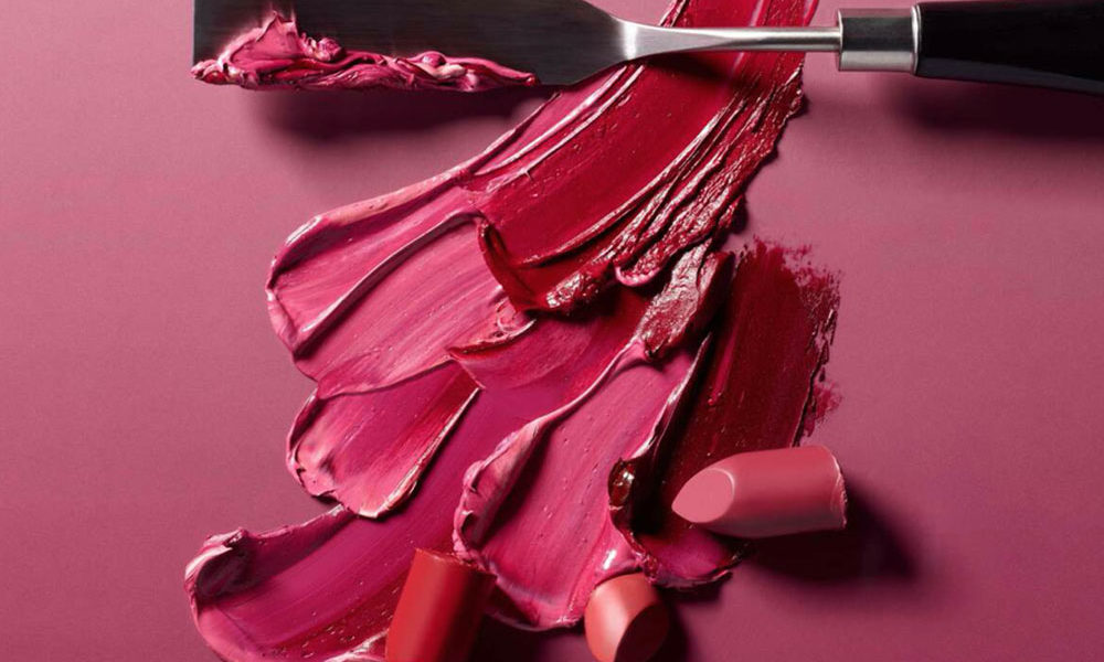 A palette of pinks, orange and red lipsticks swatched with a metal spatula.