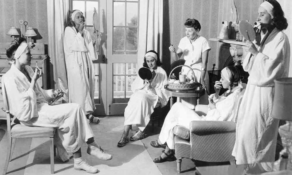 A vintage photo of women at the Elizabeth Arden spa in 1952, lounging and looking at themselves in mirrors as they apply lipstick