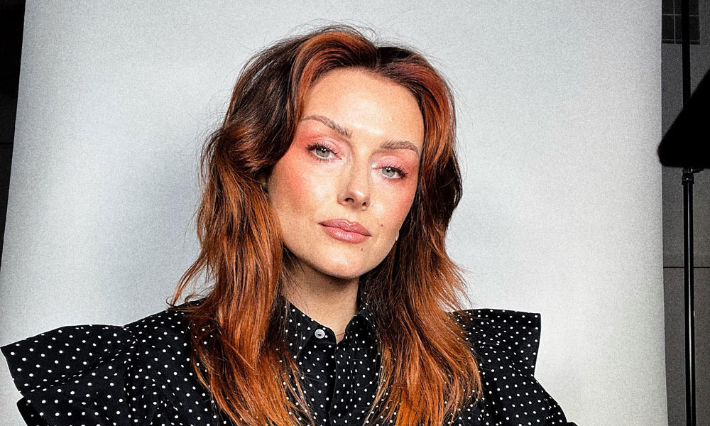 Makeup artist Katie Jane Hughes is on set with a paper backdrop behind her.