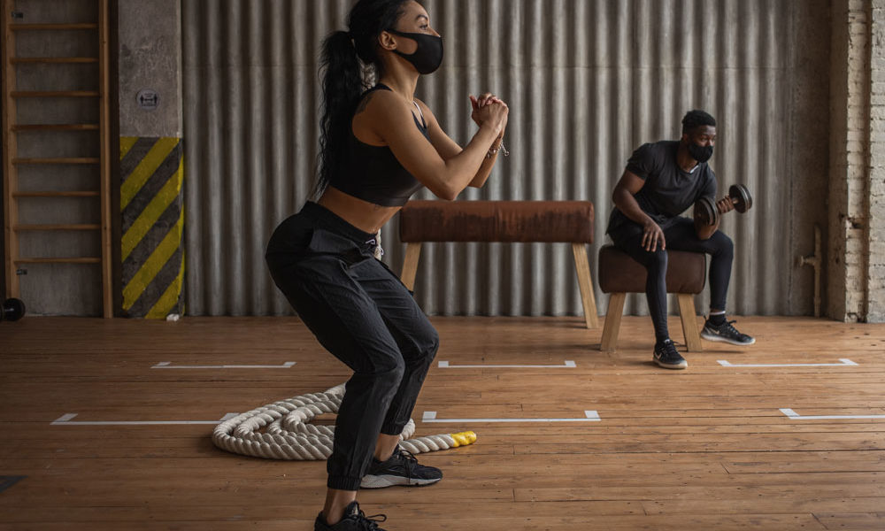 A woman working out in a gym setting wearing a face mask. She's doing squats, while another patron is doing seated curls.