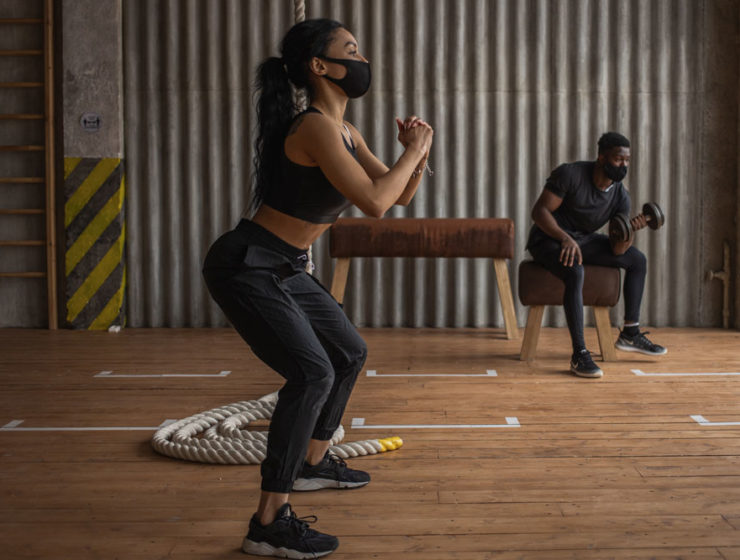A woman working out in a gym setting wearing a face mask. She's doing squats, while another patron is doing seated curls.