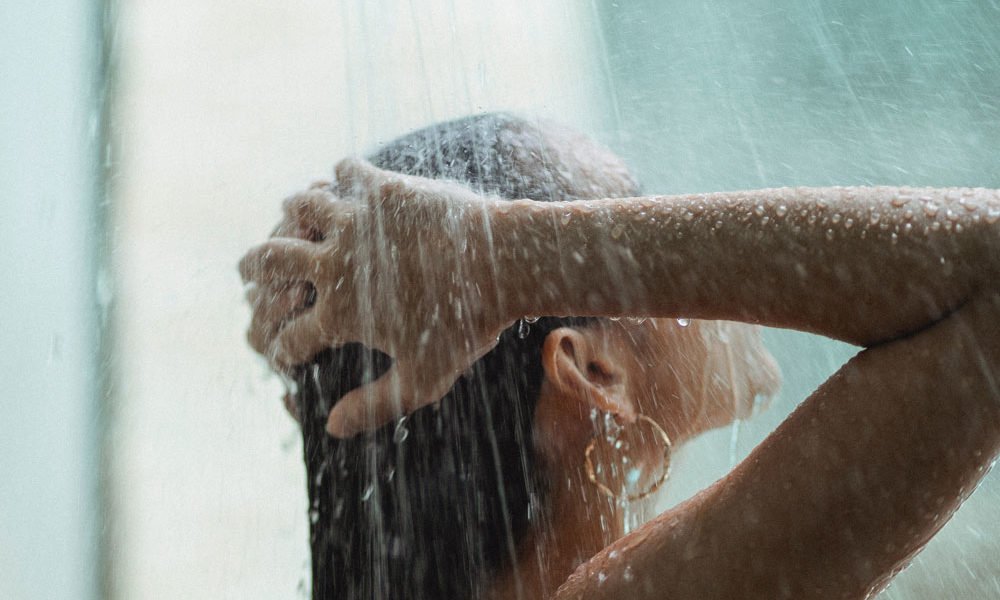 A woman enjoying a shower, with her hands above her head and large droplets of water on her hair, hands and arms