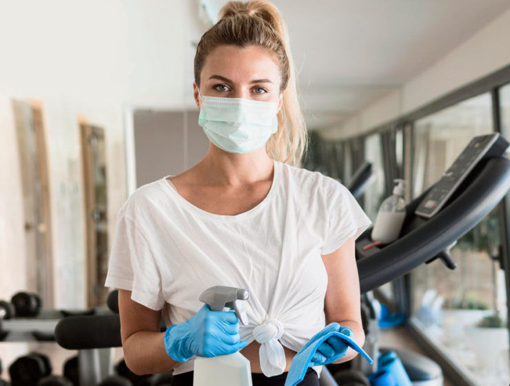 A woman in her fancy home gym, right after working out, wearing her workout clothes, a face mask, rubber gloves, while holding a spray bottle and a cloth
