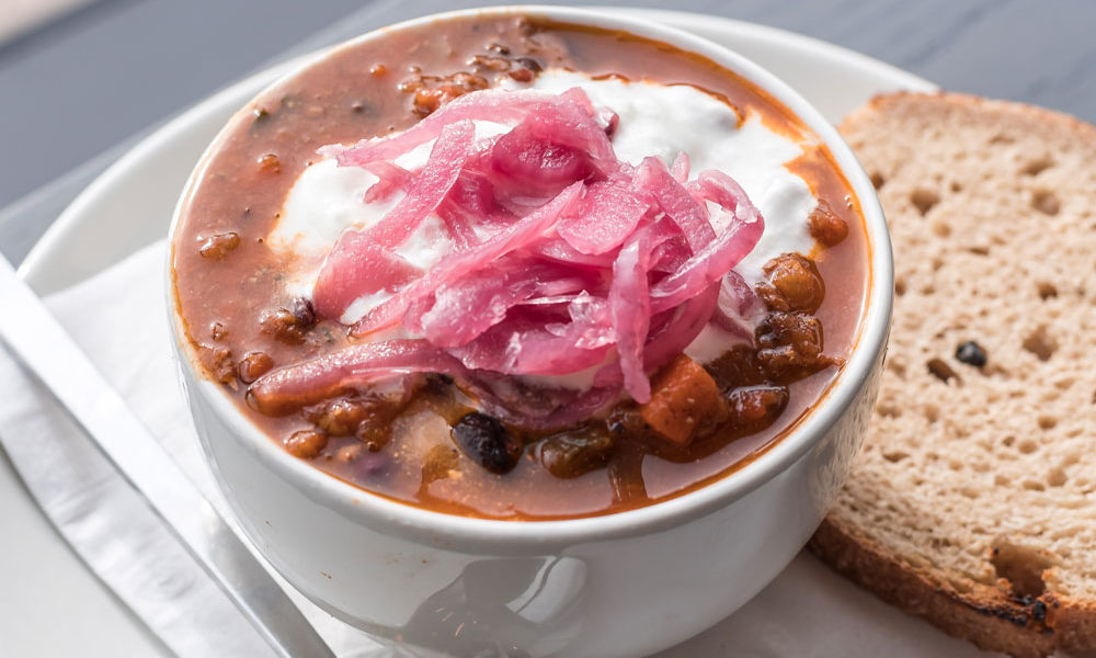 A hearty bowl of turkey chili loaded with beans and topped with pickled onions, served with a piece of rye bread.