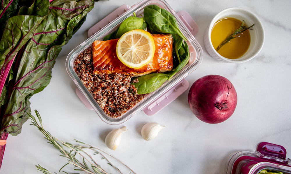 A glass dish with spinach and quinoa topped with salmon sits on a marble counter, surrounded with other ingredients.