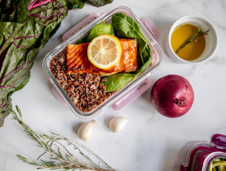 A glass dish with spinach and quinoa topped with salmon sits on a marble counter, surrounded with other ingredients.