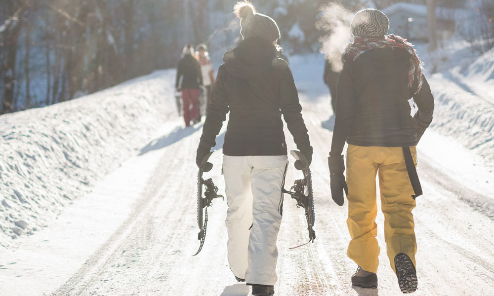 Two women are walking along a path, dressed warm, holding snowshoes, as the sun beams down on the scene.