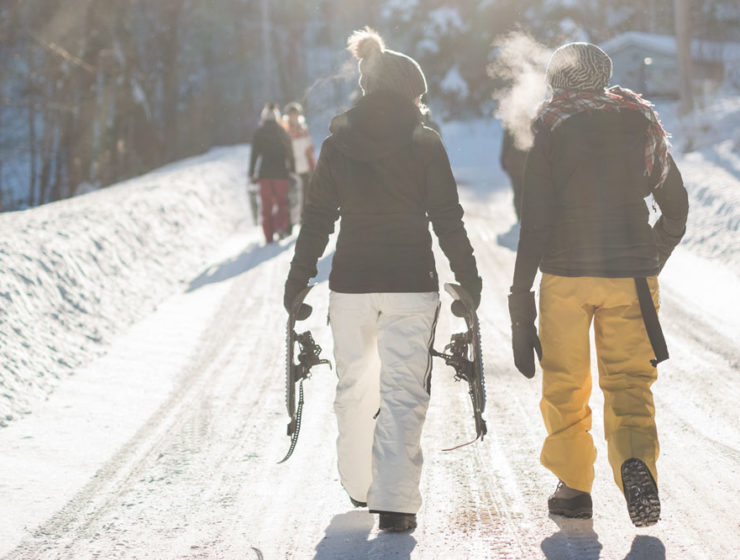 Two women are walking along a path, dressed warm, holding snowshoes, as the sun beams down on the scene.