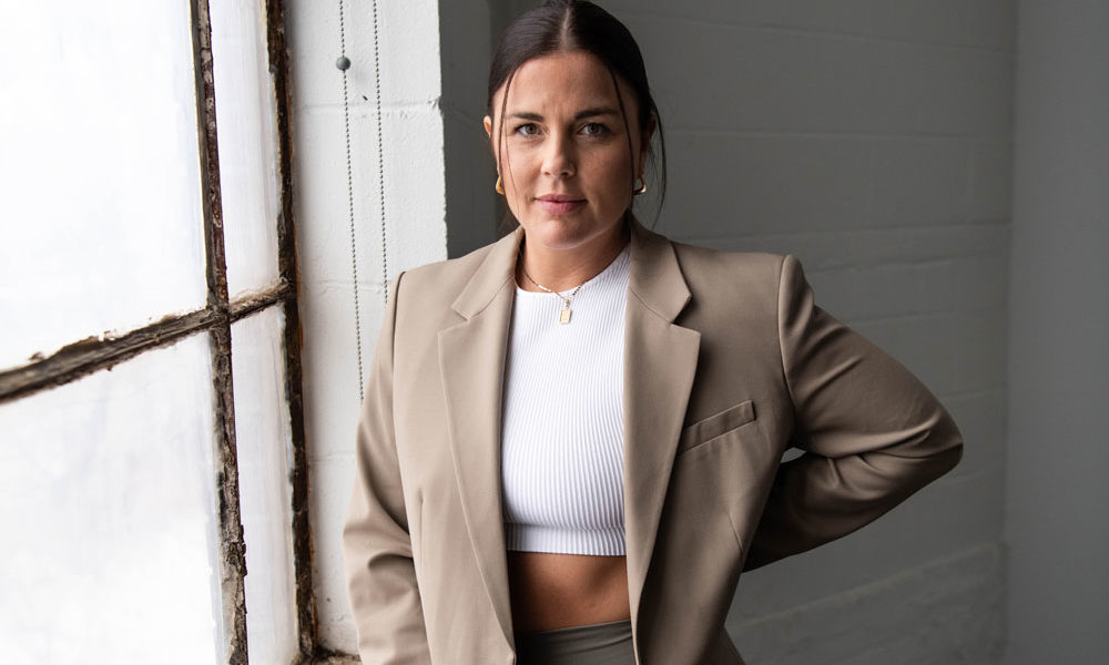 Azur Fit found Erin Ward-Williams is wearing a sports bra, leggings and a blazer, as she leans against the sill of a large warehouse window