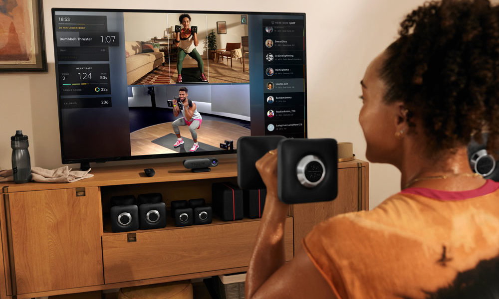 A woman is doing a squat in front of her TV, as she watches hrself and the Peloton instructor doing the same move