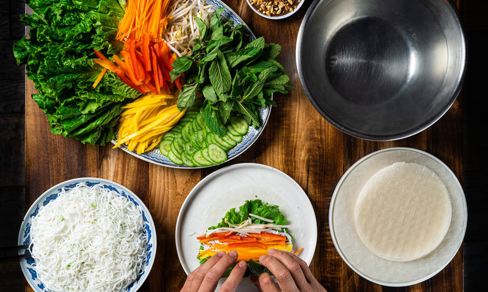 A plate full of chopped carrots, peppers, soy beans and herbs, as well as a bowl of noodles and a plate of rice wraps. A pair of hands is rolling a spring roll, too.