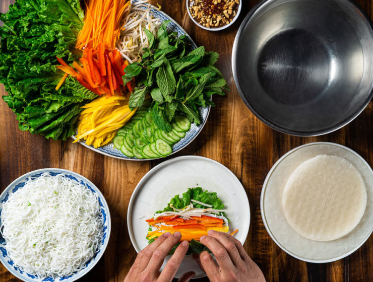 A plate full of chopped carrots, peppers, soy beans and herbs, as well as a bowl of noodles and a plate of rice wraps. A pair of hands is rolling a spring roll, too.