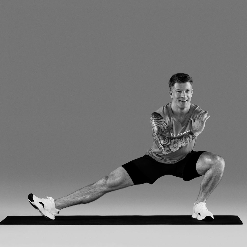 Logan Aldridge in a black and white photo demonstrates a side squat, with his left leg bent (thigh parallel to the ground) and his right leg stretched out to the side. He has his right arm crossed across his chest to touch the left shoulder, which is the arm he lost in a wakeboarding accident.