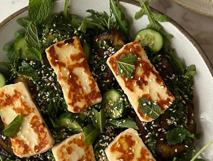 Pieces of grilled halloumi sit on a bed of cucumber and eggplant and arugula salad, sprinkled with sesame seeds, glistening with oil.